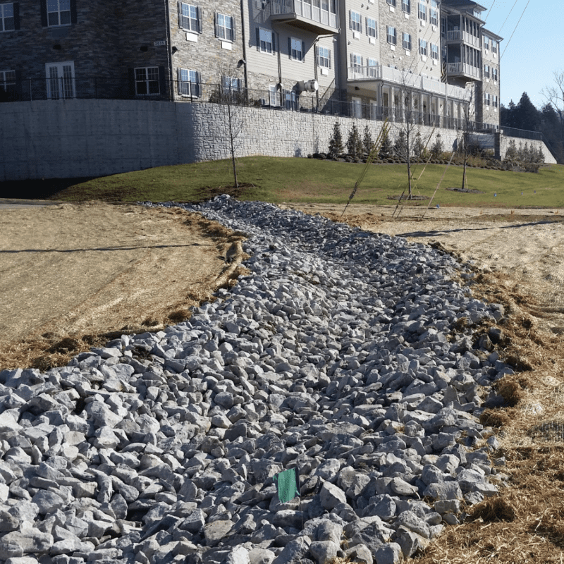 A swale lined with large gray rocks runs through a grassy area near an apartment building, designed for stormwater runoff management. This feature highlights expert grading and leveling and site preparation, with young trees by a concrete retaining wall in the background. Bushcore Excavation Drainage Site Prep Grading Middle Tennessee Dickson Nashville