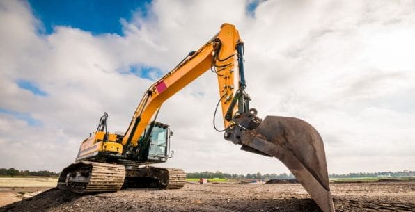 A large yellow excavator with a digging bucket is parked on a construction site, ready for excavation services. The area features dirt and gravel under a partly cloudy sky, with an open, flat landscape in the background. Bushcore Excavation Drainage Site Prep Grading Middle Tennessee Dickson Nashville
