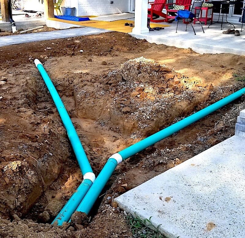 Three large blue pipes are laid in a trench by a sitework contractor next to a house under construction, surrounded by exposed soil, stacked bricks, and newly installed paving stones. Bushcore Excavation Drainage Site Prep Grading Middle Tennessee Dickson Nashville