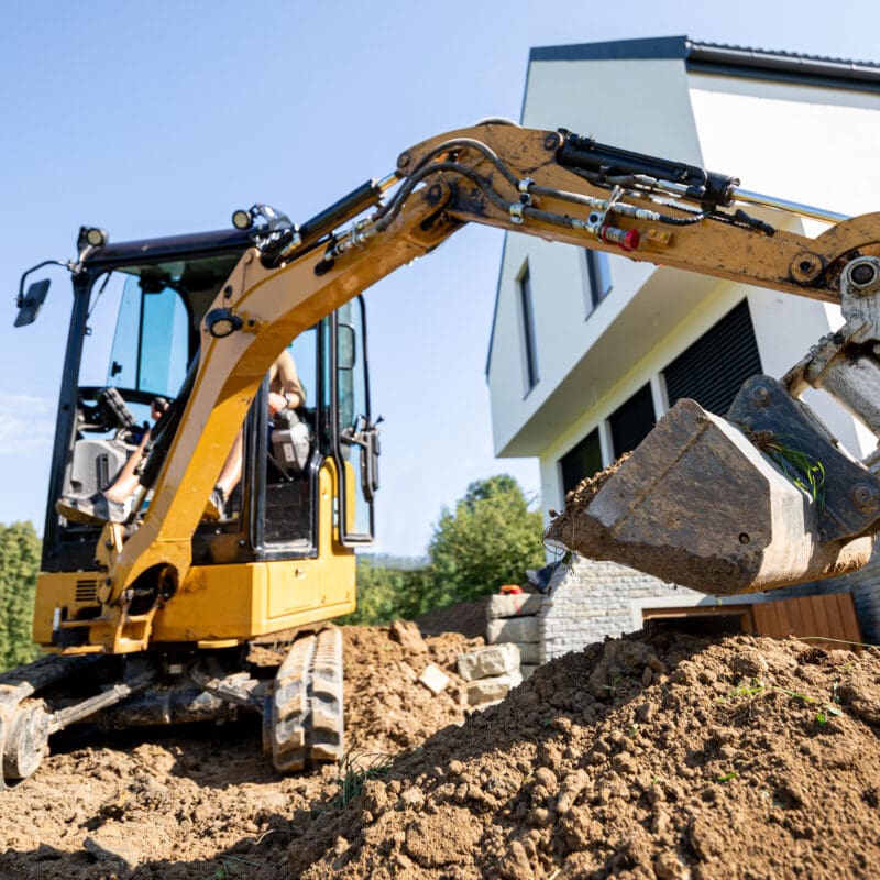 A yellow excavator performs grading and leveling in front of a modern white house under construction, with trees and greenery in the background on a sunny day. Bushcore Excavation Drainage Site Prep Grading Middle Tennessee Dickson Nashville