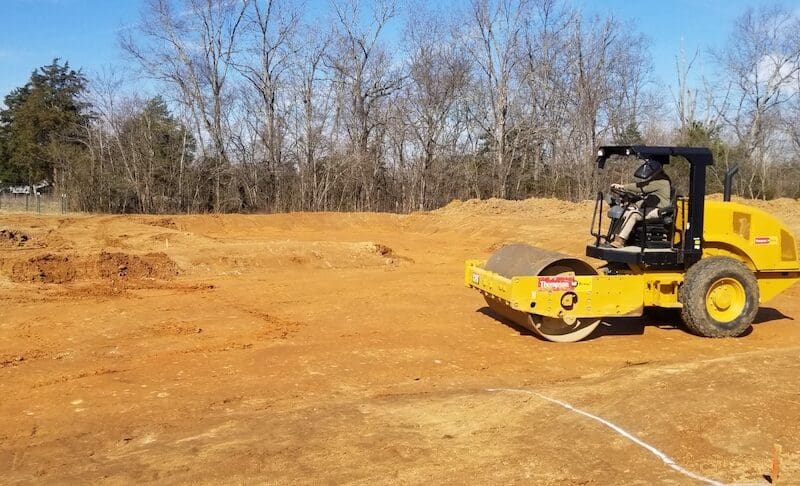 A yellow steamroller compactor is parked on a cleared construction site, highlighting site preparation and grading and leveling work, with trees and a blue sky in the background. Bushcore Excavation Drainage Site Prep Grading Middle Tennessee Dickson Nashville