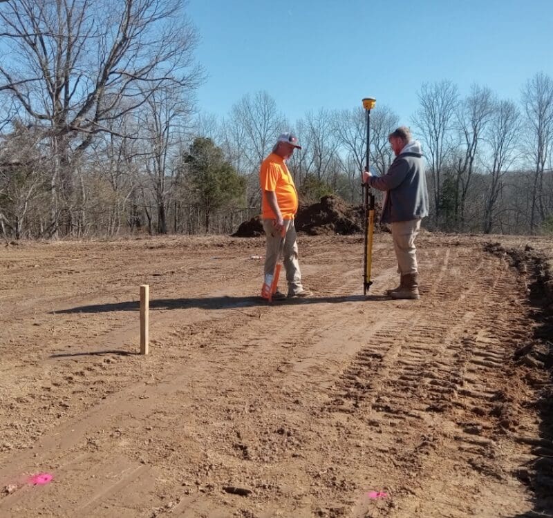 Two men stand on a leveled dirt construction site using surveying equipment, surrounded by bare trees and wooden stakes. A sitework contractor oversees excavation services under a clear, sunny sky. Bushcore Excavation Drainage Site Prep Grading Middle Tennessee Dickson Nashville
